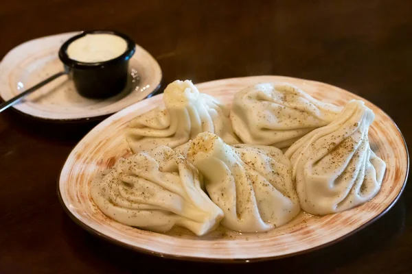 Khinkali - traditional Georgian dumplings in a white plate with white sour cream sauce on a wooden table in the restaurant — 图库照片
