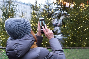 Tanınmayan bir kadın cep telefonuyla Noel ağacının fotoğrafını çekiyor. Arkadan insanlar.