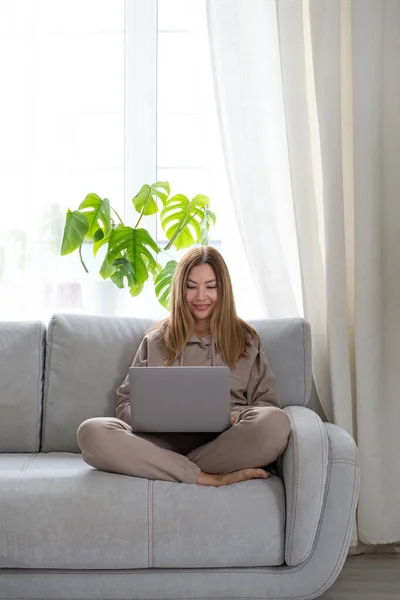 Fiatal vagy középkorú nő ül laptoppal szürke kanapén az otthoni irodában. A távoli munkahely fogalma és az otthoni munkavégzés. — Stock Fotó