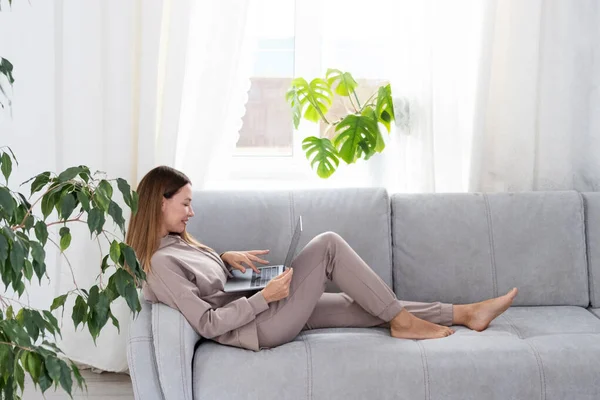 Young or middle age woman sitting with laptop on grey couch in home office with monstera plant. Concept of remote workplace and working at home. Stock Photo