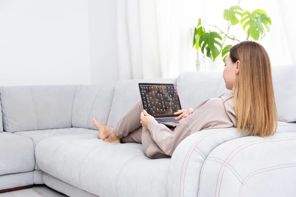 Mulher jovem ou de meia idade sentada com laptop no sofá cinza no escritório em casa com planta monstera. Conceito de local de trabalho remoto e trabalho em casa. Imagens De Bancos De Imagens Sem Royalties