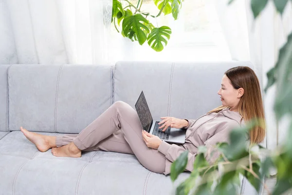 Fiatal vagy középkorú nő ül laptoppal szürke kanapén otthoni irodában monstera növény. A távoli munkahely fogalma és az otthoni munkavégzés. — Stock Fotó