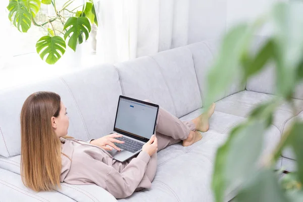 Fiatal vagy középkorú nő ül laptoppal szürke kanapén otthoni irodában monstera növény. A távoli munkahely fogalma és az otthoni munkavégzés. — Stock Fotó