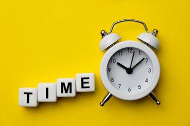 White analog alarm clock isolated on yellow background with word Time on white plastic cubes. Concept of Time. A Clock with the word TIME. Copy space. Close-up.