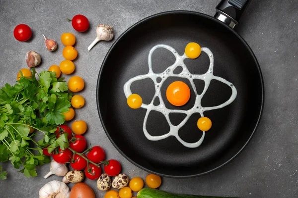 黒いフライパンに鶏やウズラの卵から原子のイメージの形で元の揚げ卵 トップビュー フラットレイアウト 灰色の背景に赤と黄色のチェリートマト ニンニクとパセリ ストック画像