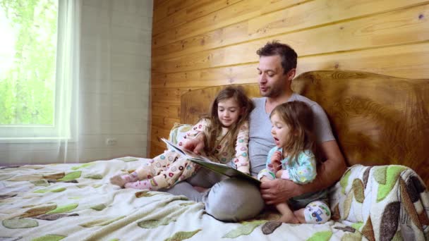 Father sits on the bed with his cute little daughtersin and reads the book. — Stock Video