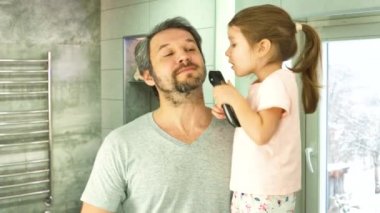 Bir kız banyodaki aynaya bakarak babalarının sakalını saç makasıyla kesiyor.
