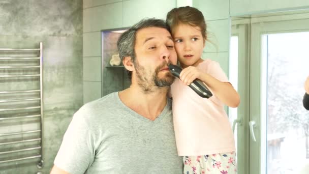 Uma menina corta a barba dos pais com um cortador de cabelo, olhando em um espelho em um banheiro — Vídeo de Stock