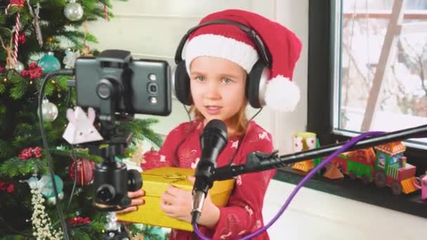 Egy lány fülhallgatóban és egy mikrofonba énekel egy karácsonyfa mellett. — Stock videók