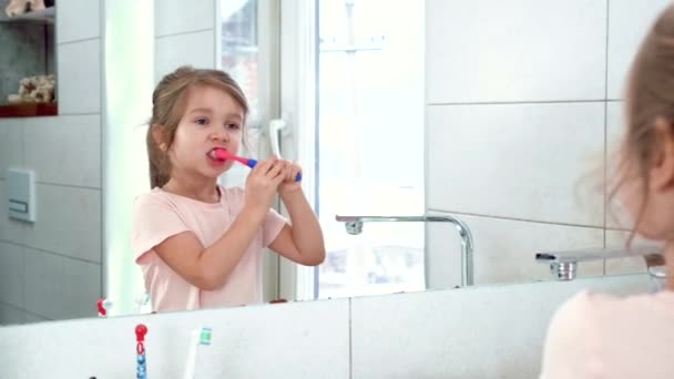 Diş fırçalı küçük kız, pencerenin yanındaki aynaya bakıyor.. — Stok video
