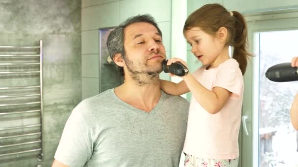 Una chica corta la barba de los papás con una cortadora de pelo, mirando en un espejo en un baño — Vídeo de stock