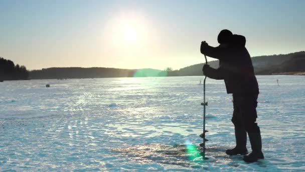 A fisherman makes a hole in the ice for winter fishing. Sunset on winter lake. — Wideo stockowe