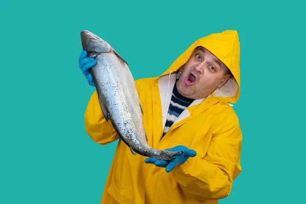 Рыбак в жёлтом костюме держит в руках свежепойманную рыбу. рыбака удивляет хороший улов. Фишер на морском фоне. Ловец в дождевике. — стоковое фото