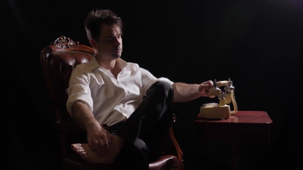 Un hombre en negro en una camisa blanca se sienta en una silla — Vídeo de stock