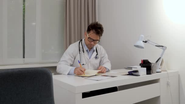 医者はホワイトオフィスのテーブルの上の処方箋や写真のデータをノートに書いて — ストック動画