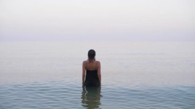 a girl in an evening dress stands in the water and then turns and looks at the camera