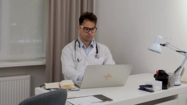 a male doctor works at a computer and writes out key treatment factors in a notebook
