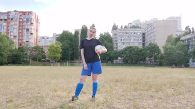 girl footballer holding the ball in her hands looks at the camera and stands in an abandoned stadium against the backdrop of residential buildings