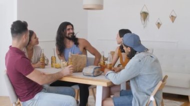 a group of 5 friends are sitting at a table, talking when suddenly one of the guys exclaims sharply and raises his hands to drink for his birthday, clinking bottles of beer