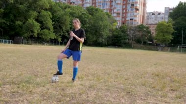 woman football player stands with her foot on the ball breathes heavily and drinks water