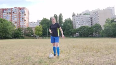 a beautiful female football player stands in an abandoned stadium between houses, puts her foot on the ball and looks at the camera smiling