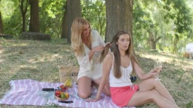 a girl on her knees twists pigtails to her friend at a picnic in the park
