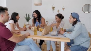 a group of 5 friends are sitting at a table, talking when suddenly one of the guys exclaims sharply and raises his hands to drink for his birthday, clinking bottles of beer