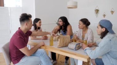 a group of 5 friends are sitting at a table, talking when suddenly one of the guys exclaims sharply and raises his hands to drink for his birthday, clinking bottles of beer