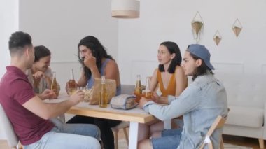 a group of 5 friends are sitting at a table, talking when suddenly one of the guys exclaims sharply and raises his hands to drink for his birthday, clinking bottles of beer