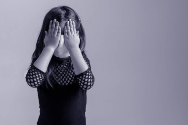Tıbbi maskeli bir kız ellerini gri bir arka planda tutarak yüzünü kapattı. Tecrit sırasında küçük üzgün çocuk