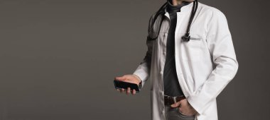 Elinde steteskop ve telefon bulunan beyaz üniformalı bir erkek doktor ya da pratisyen hekimin yakın çekimi kaliteli tıbbi bakım gösteriyor. Beyaz önlüklü, fonendoskoplu bir doktor gri arka planda duruyor. sağlık hizmeti kavramı.