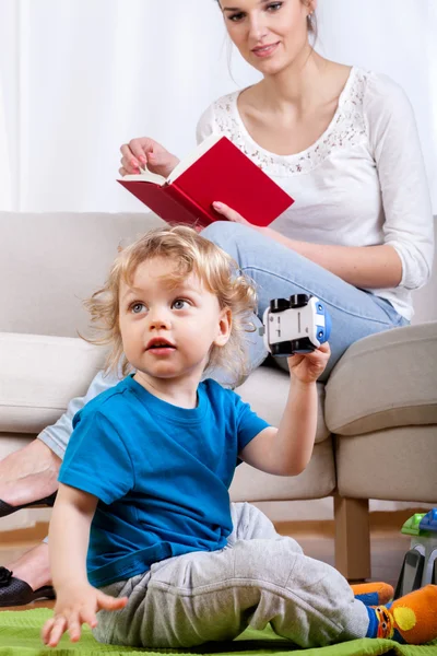 Niño jugando mientras la madre está leyendo — Foto de Stock