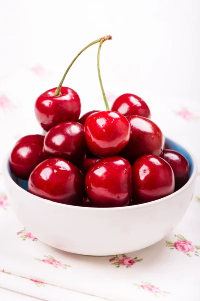 Primer plano de cerezas rojas con gotas de agua en tazón blanco en servilleta con rosas en la mesa — Foto de Stock