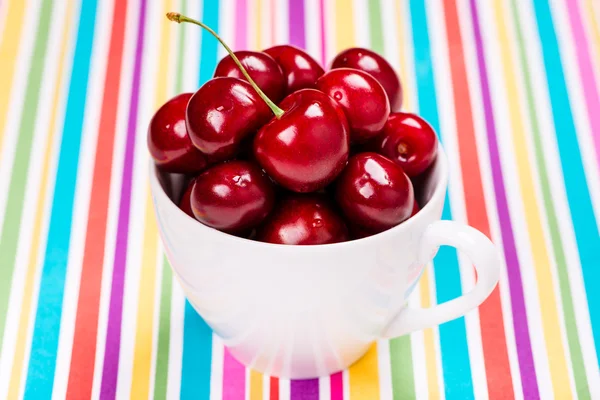 Nahaufnahme von roten Kirschen mit Wassertropfen in weißer Tasse auf buntem Hintergrund — Stockfoto