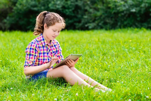 Venkovní portrét krásné dospívající dívky v neformálním oblečení sedí v trávě s digitálním tabletu na kolena, čtení a procházení — Stock fotografie