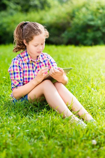 Retrato al aire libre de una bonita adolescente vestida con ropa casual sentada en el césped con una tableta digital en las rodillas, leyendo y surfeando —  Fotos de Stock