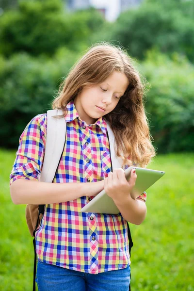 Outdoor portrait of a beautiful teenager girl in casual clothes with backpack holding digital tablet in her hand, typing and reading — Stock Photo, Image