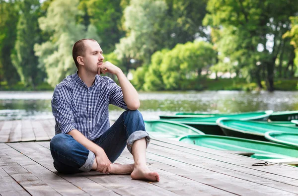 Joven hombre barbudo guapo sentado en el muelle de madera en el día de verano, relajante y pensando — Foto de Stock