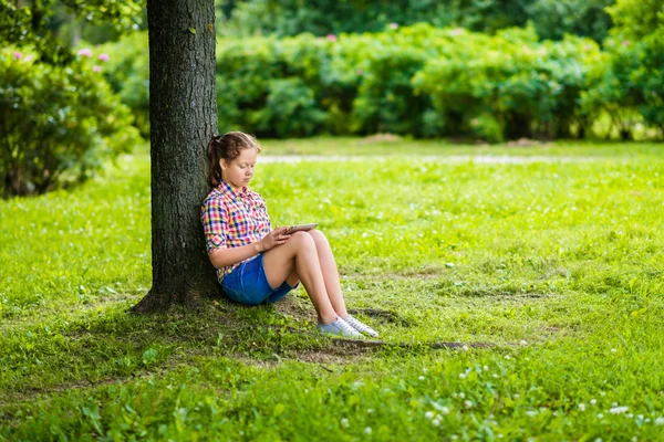 Linda adolescente en ropa casual con tableta digital en sus rodillas en el parque bajo el árbol — Foto de Stock