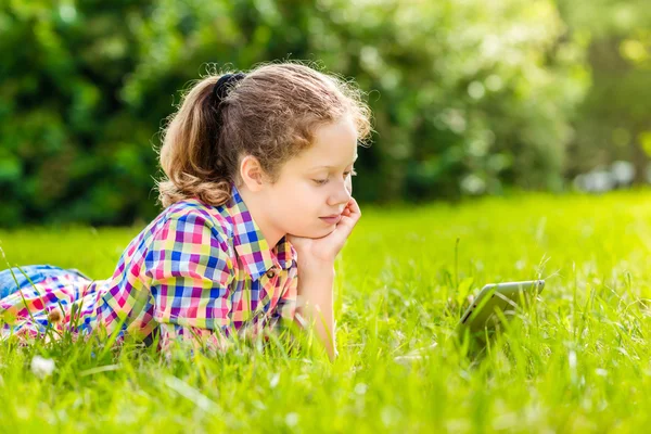 Hermosa adolescente con ropa casual acostada en la hierba a la luz del sol con tableta digital o libro electrónico, retrato al aire libre —  Fotos de Stock