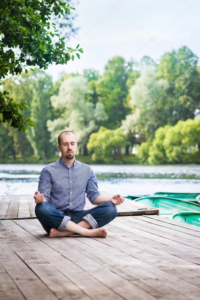 Young attractive breaded man with closed eyes sitting on wooden pier, relaxing and meditating in summer morning