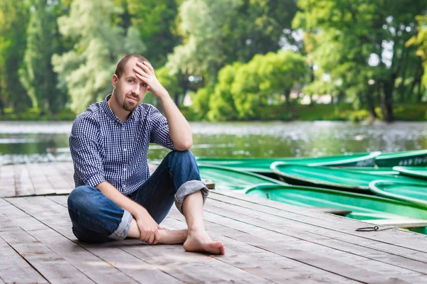 Joven hombre barbudo guapo sentado en el muelle de madera en el día de verano y sonriendo — Foto de Stock