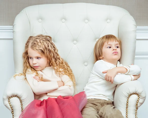 Irmãozinhos bonitos (menino e menina) estando em desacordo uns com os outros, sentados em poltrona Imagens De Bancos De Imagens Sem Royalties