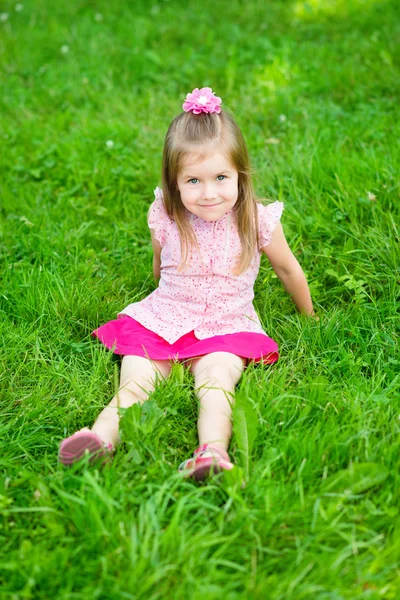 Menina bonito com longos cabelos loiros em blusa rosa e saia sentado na grama no parque de verão, retrato ao ar livre — Fotografia de Stock