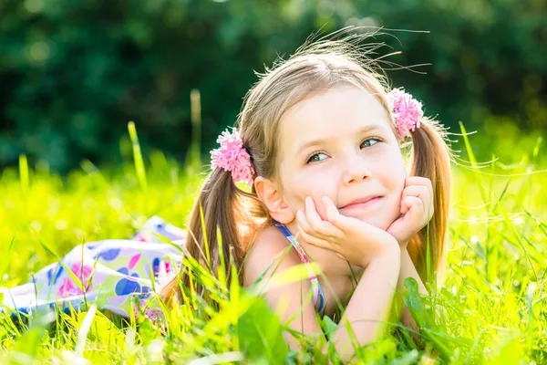Cute smiling little girl with two blond ponytails laying on grass in summer park, outdoor portrait Royalty Free Stock Photos