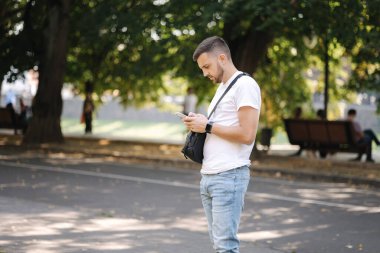 Beyaz tişörtlü, siyah çantalı bir adam telefon Outddor kullanıyor. Adamın akıllı saati var.
