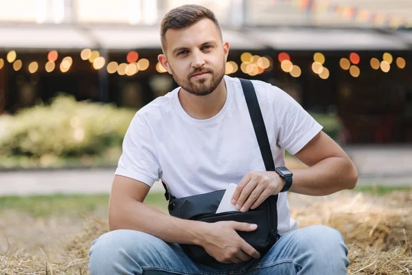 Hombre guapo tomar el teléfono de la bolsa de hombro. El hombre se sienta en el banco de heno al aire libre — Foto de Stock