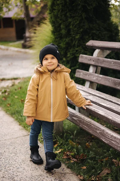 재킷을 입고 모자를 쓴 행복하고 귀여운 소녀가 벤치 옆 야외의 사진 작가에게 포즈를 취한다. 작은 모델은 가을에 재밌게 놀아 — 스톡 사진