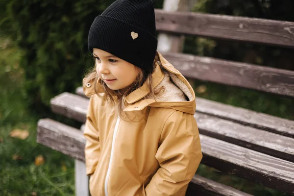 재킷을 입고 모자를 쓴 행복하고 귀여운 소녀가 벤치 옆 야외의 사진 작가에게 포즈를 취한다. 작은 모델은 가을에 재밌게 놀아 — 스톡 사진