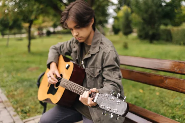 Adolescente bonito tocando guitarra acústica ao ar livre no Outono. Menino sentado no banco e tocando música — Fotografia de Stock